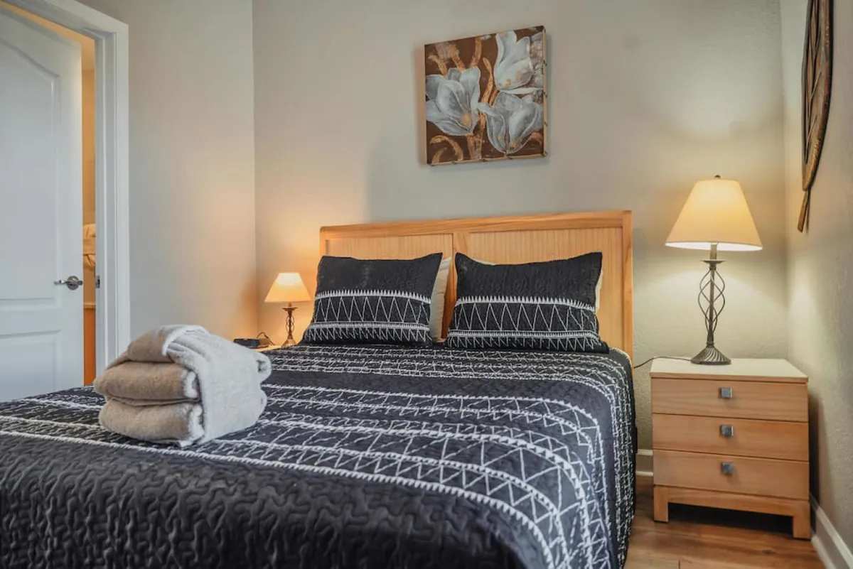 A peaceful bedroom showcasing a comfortable bed, decorative artwork above the headboard, lamps on the nightstand, and a bedside table. The subdued color palette and thoughtful details create a sense of quiet retreat.