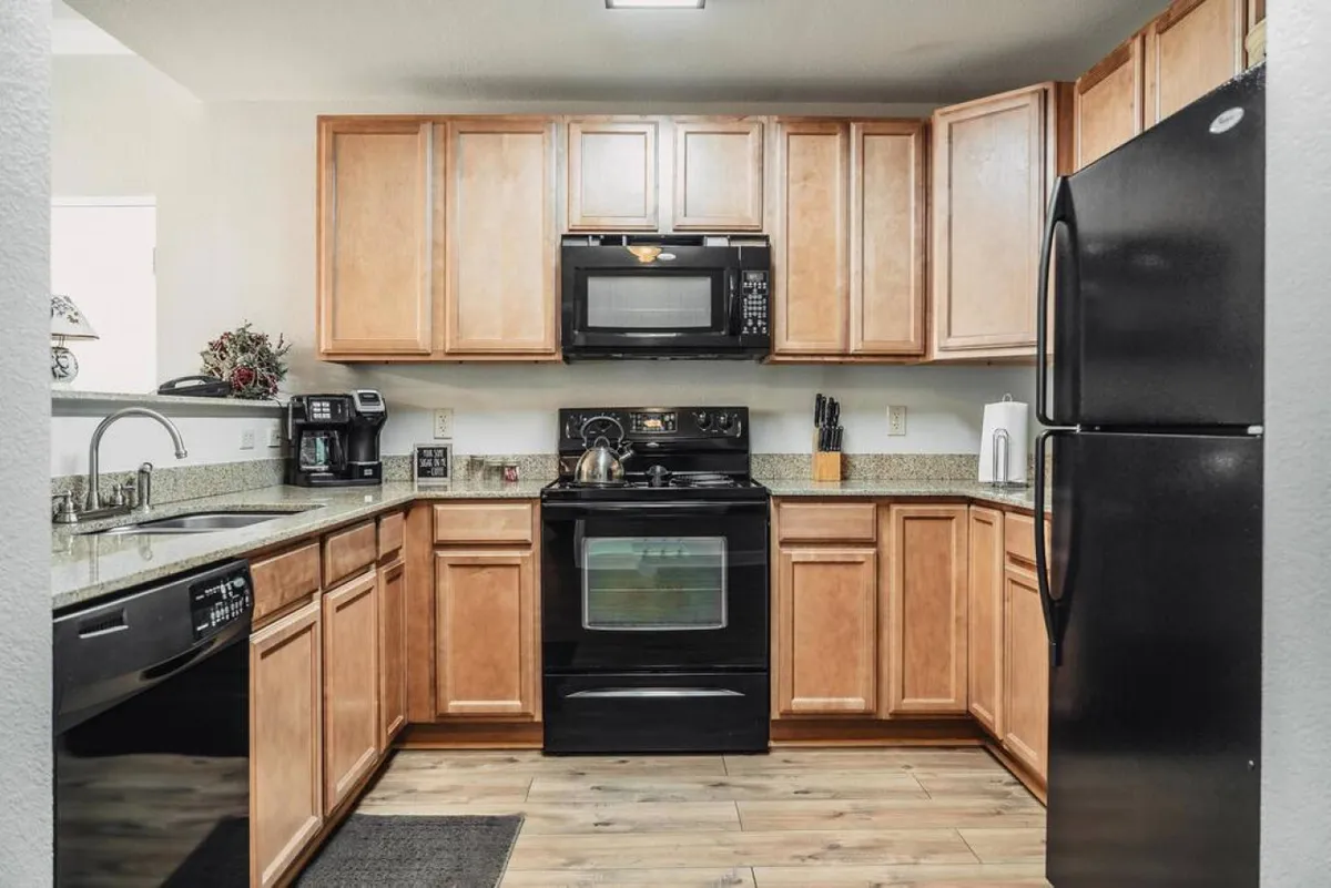 A bright kitchen featuring black appliances, granite countertops, wooden cabinetry, and a kitchen sink. The space is staged for meal preparation, with clean, modern surfaces and an inviting overall look.