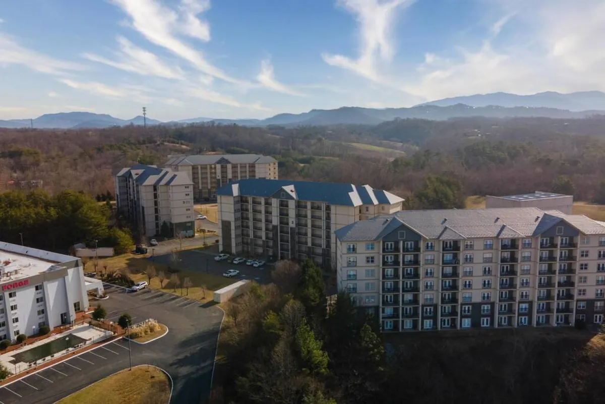 Multi-story residential buildings nestled in a landscaped area with open green spaces and a scenic mountain backdrop. The layout includes generous sidewalks and gathering spaces where residents can walk, socialize, or relax, all within a barrier-free outdoor environment.