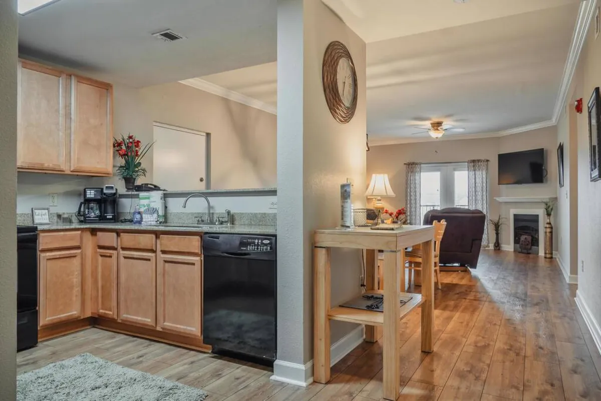 A wide, inviting kitchen and dining area with kitchen appliances, a wooden dining table, a comfortable seating area, and decorative elements like plants and a clock. The scene suggests social cooking and group dining.