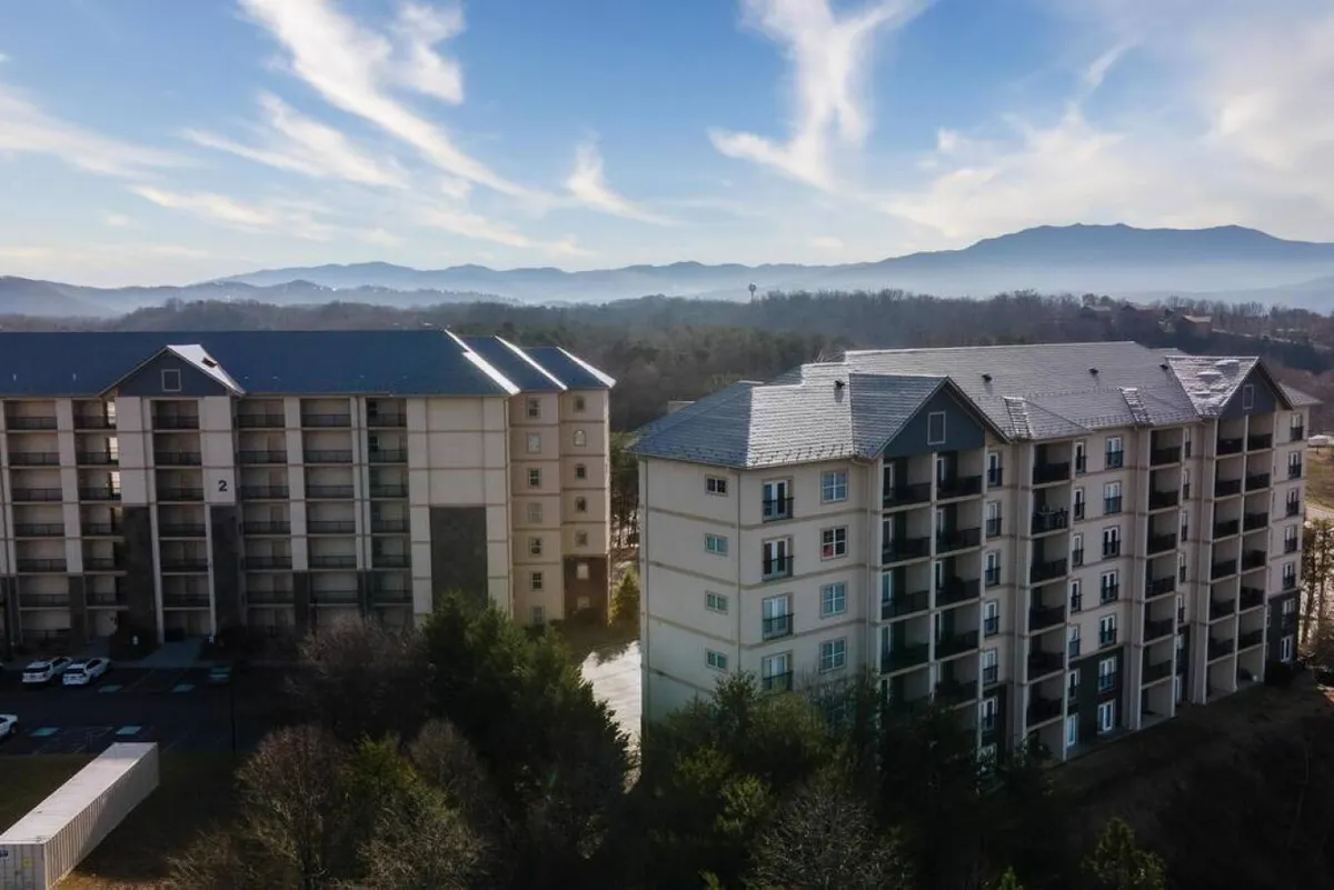 This image shows two apartment buildings set against a backdrop of distant mountains, with a blue sky adorned with wispy clouds above. The peaceful residential setting is highlighted by the spacious outdoor area and unobstructed views of the mountains.