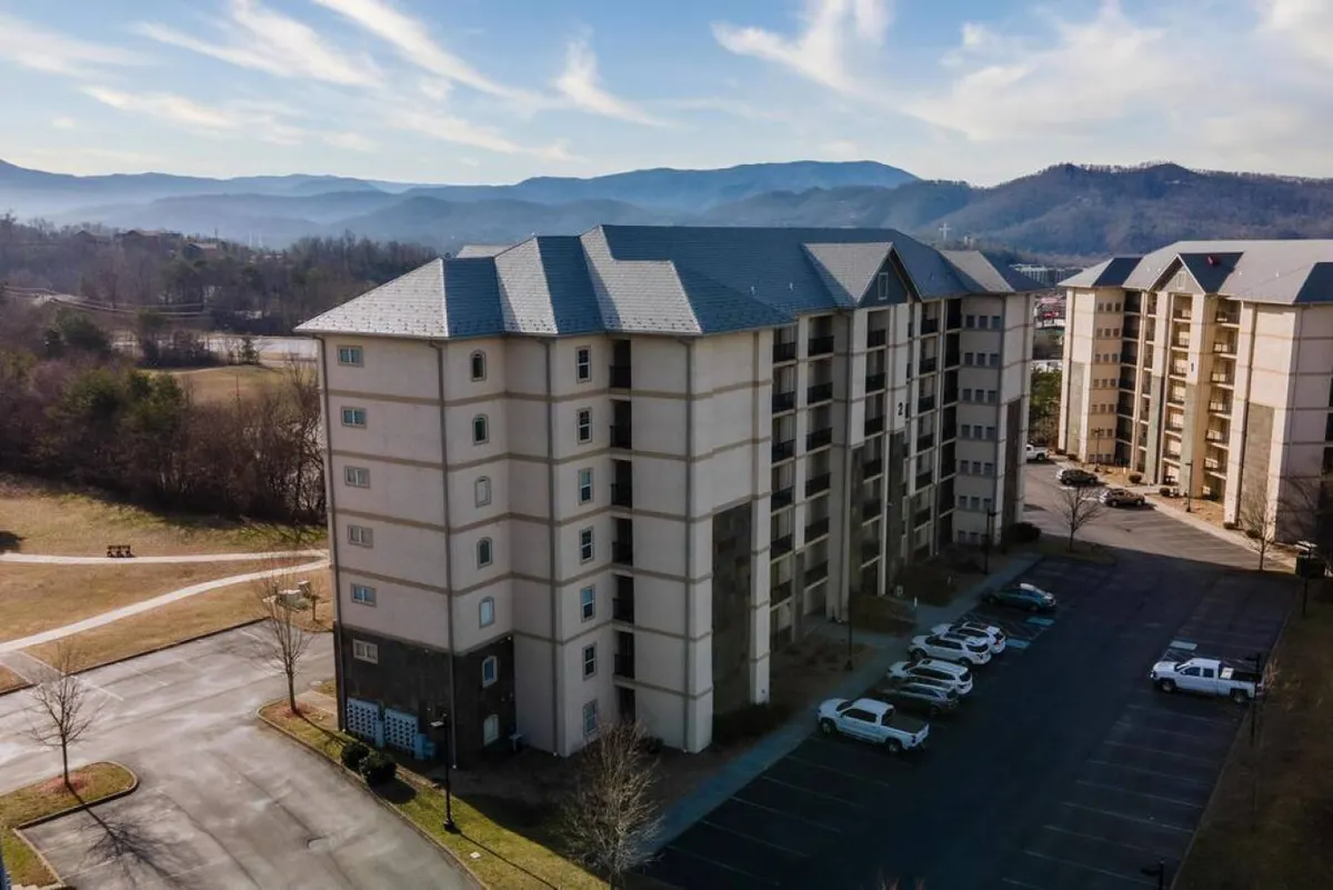 This image features a multi-story building nestled against beautiful surrounding mountains, with a spacious parking lot in the foreground. The serene setting emphasizes both the scale of the accommodation and its integration with the natural landscape of Pigeon Forge.