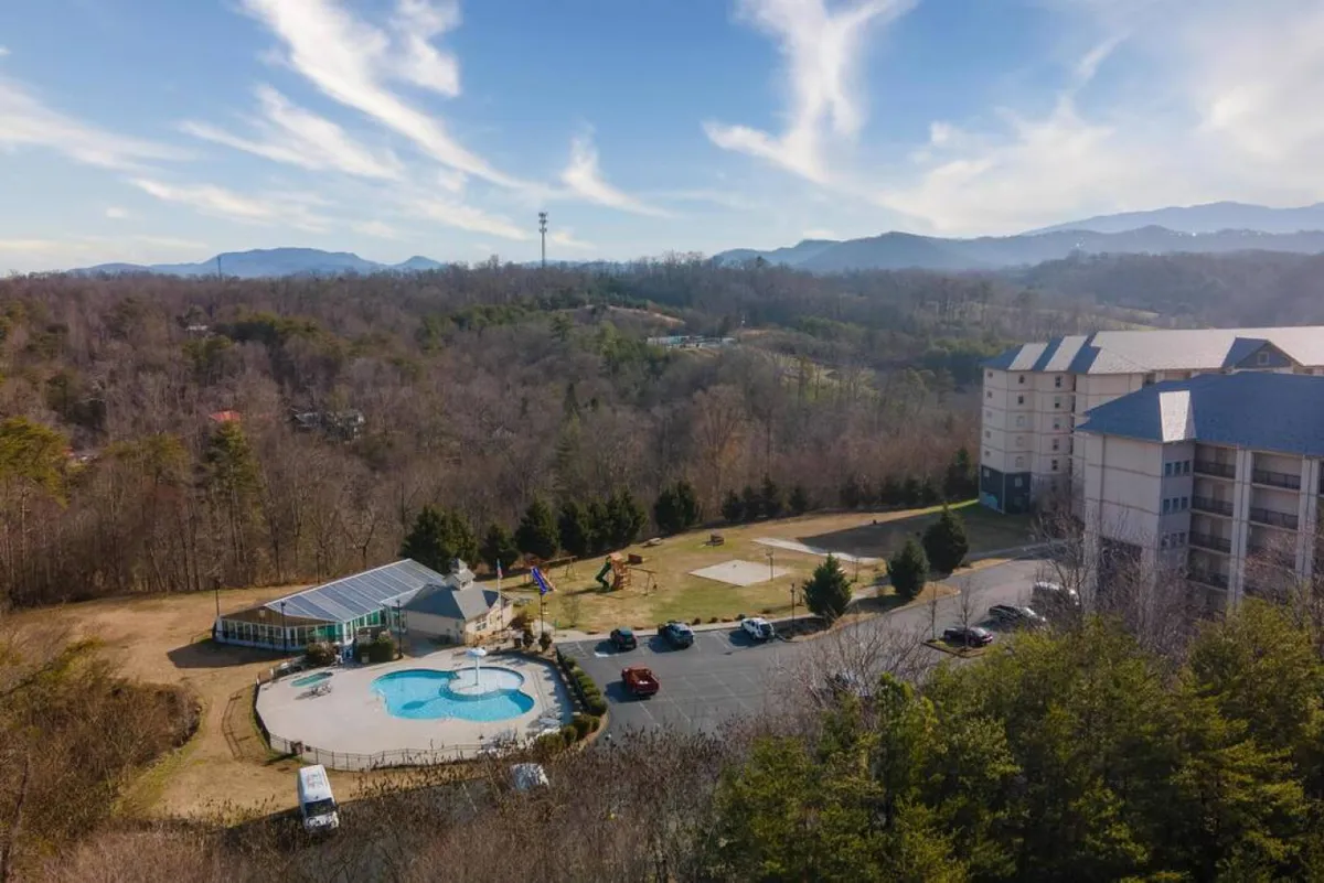 This image captures the vibrant resort outdoor area, featuring a swimming pool, a playground for children, lush green spaces, and the dramatic backdrop of the surrounding mountains. The hotel building stands nearby, situating the amenities in a resort context.