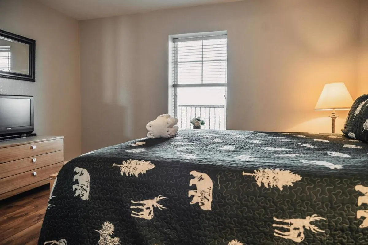 A cozy bedroom with a patterned quilt on the bed, a nightstand with a lamp, a television mounted on the wall, and a mirror. Soft natural light streams in, and the room feels inviting and comfortable, perfect for unwinding at any time of day.