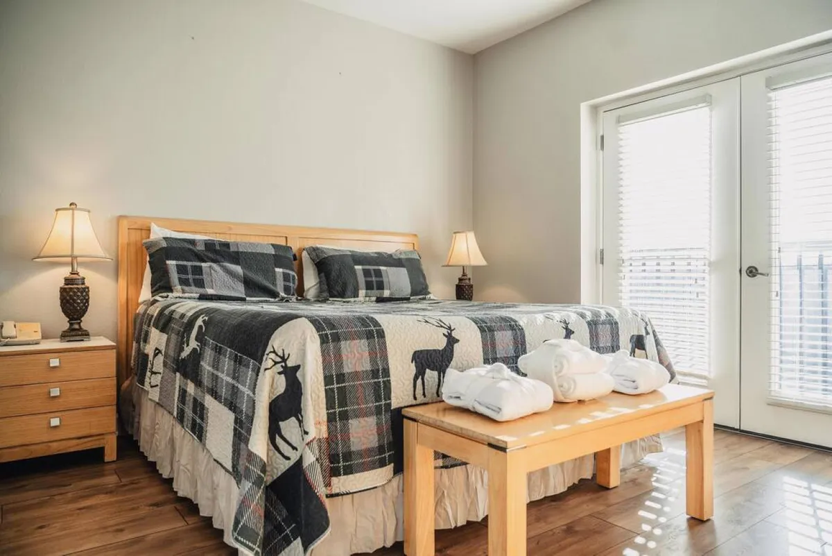 The image showcases a warm, inviting bedroom featuring a comfortable bed adorned with a patterned quilt, flanked by cozy nightstands and softly glowing lamps. Sliding glass doors fill the space with natural light and hint at a beautiful outdoor view, creating an atmosphere perfect for relaxation and peaceful mornings.