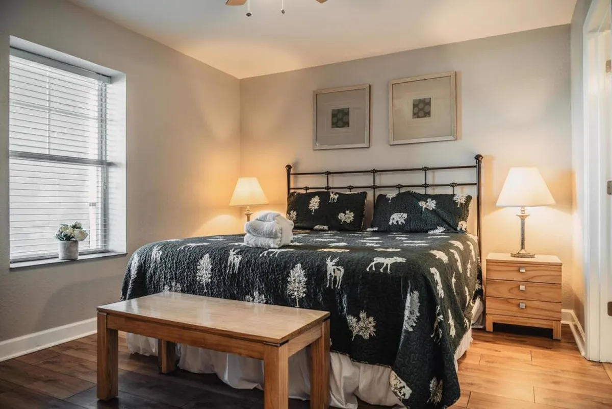A serene bedroom featuring a large bed with a decorative blanket, nightstands with lamps on either side, a wooden bench at the foot of the bed, and a window allowing natural light to filter in. The room exudes a tranquil and inviting atmosphere, perfect for rest and relaxation.
