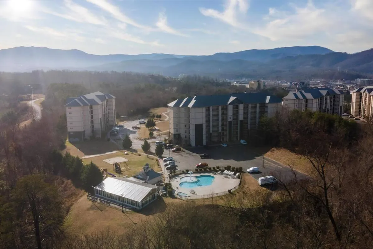 This image captures a vibrant outdoor swimming pool surrounded by mountains, residential buildings, and landscaped communal areas. The scene is bright and inviting, suggesting a lively atmosphere for family fun and relaxation. People are seen enjoying poolside activities, reinforcing the idea of community and leisure.