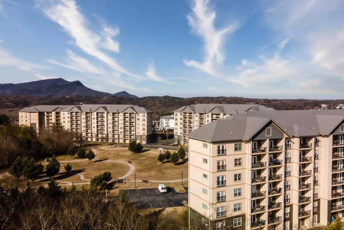 A peaceful landscaped area with green lawns, winding walking paths, mountain views in the background, and apartment buildings that hint at the accessibility and connection between accommodations and outdoor amenities.