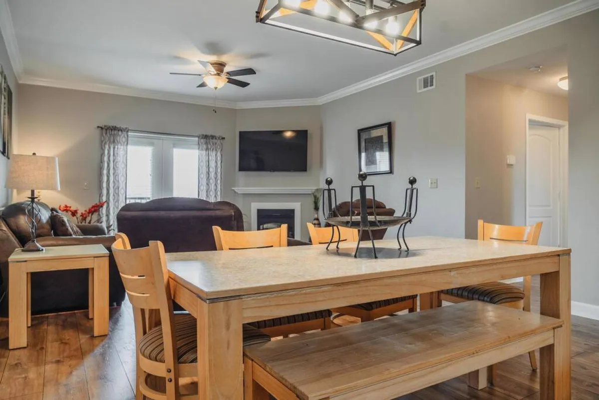 A cozy, open-plan living and dining area featuring a dining table set for a meal, a plush sofa, a glowing fireplace, and a prominent flat-screen television. The space is warmly lit and arranged for both relaxation and group gatherings, with ample room for family and friends to enjoy each other's company.