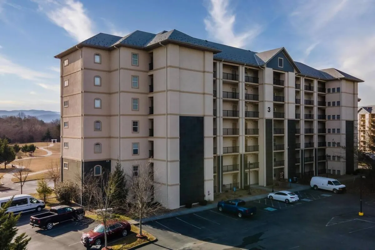 A four-story building with wide paved walkways and a visible parking area, surrounded by mature trees and green landscaping. The broad sidewalks and unobstructed entry make it easy for guests with mobility needs to access the building. The overall setting appears serene and inviting, with ample outdoor space for movement and relaxation.