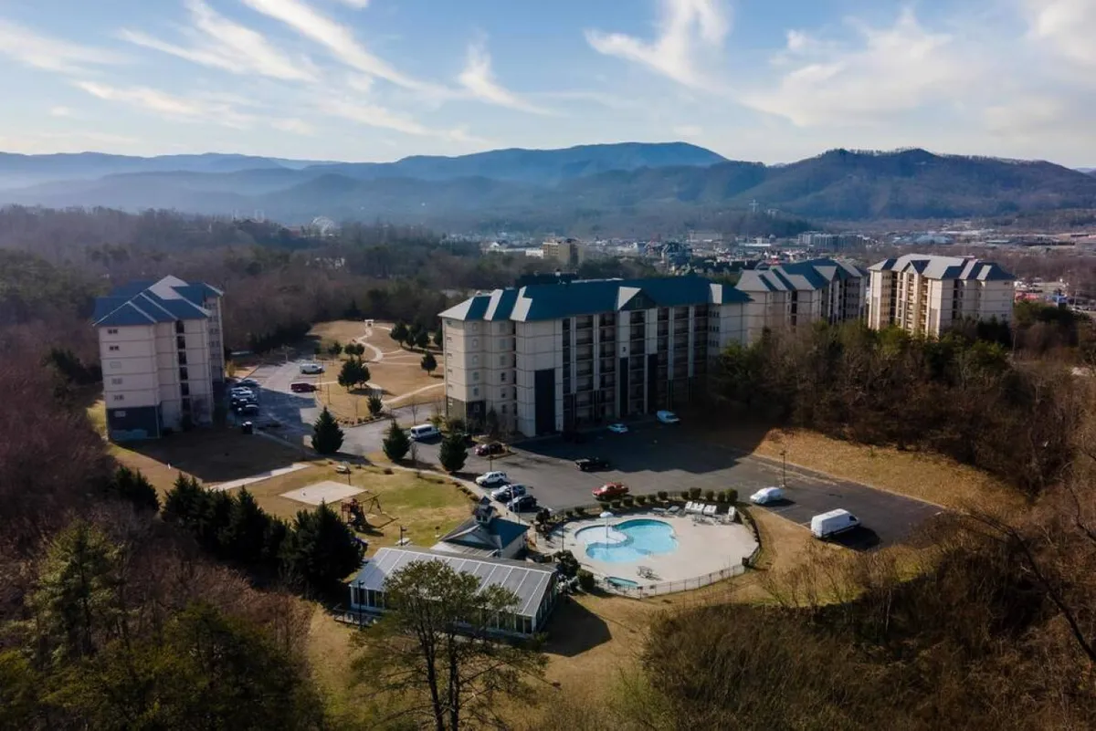 The resort’s swimming pool sits against a backdrop of rolling mountains, with resort buildings and a spacious parking area nearby. The scene is vibrant yet tranquil, suggesting both fun and relaxation for groups.