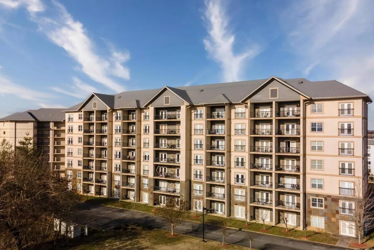 The image displays the exterior of a modern apartment building with balconies and a landscaped area below. The sunset-lit scene conveys a welcoming, family-friendly atmosphere, and showcases both the comfortable accommodations and community vibe.
